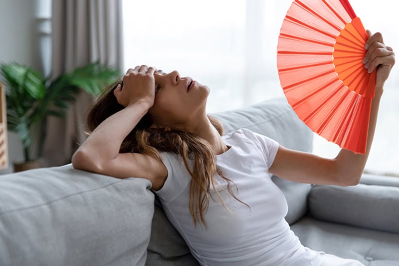 Why Is My AC Blowing Hot Air? Photo of a young woman fanning herself on her couch to keep cool.